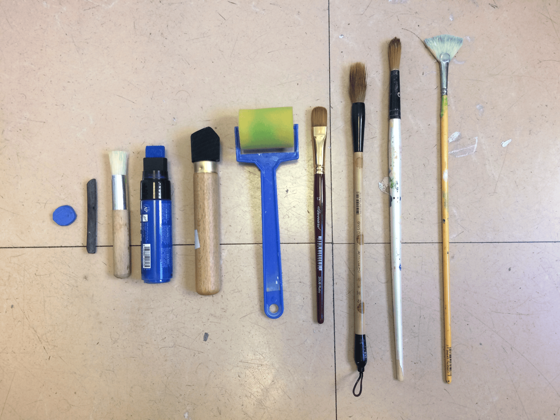 An image of various tools sorted from smallest to biggest. From left to right: tailor’s chalk, carbon, brush, Posca pen, carbon mounted to a stick, soft roller, brush, chinese brush, fine brush, wide brush.