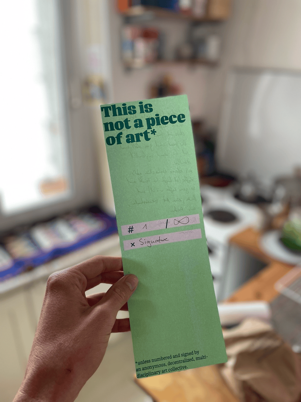 A photo of my hand holding a green, riso-printed piece of art. It reads: »This is not a piece of art*«. In the center there is room to be signed and numbered. At the bottom it reads: »*unless numbered and signed by an anonymous, decentralized, multidisciplinary art collective.«.